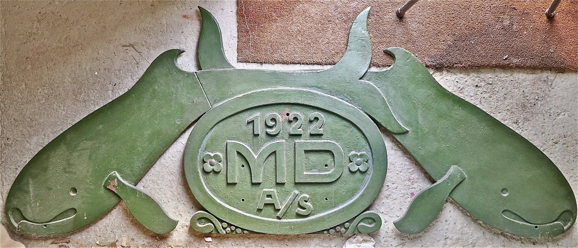 Grønmalet logo fra 1922 med to stiliserede marsvin og teksten MD A/S